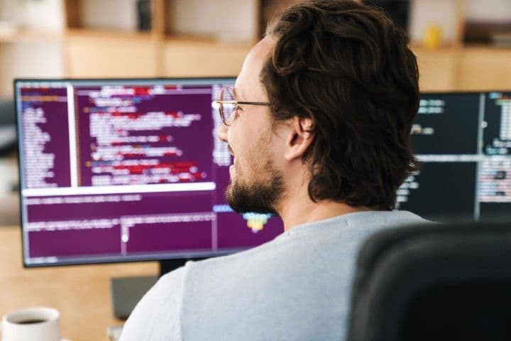Développeur vue de dos à son bureau. On voit ses écrans ouvert sur des logiciels de code.